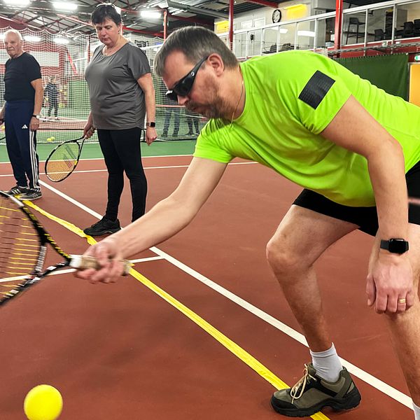 En tennisövning på inomhustennisbana där en synskadad spelare i grönt tränar sin teknik genom att slå en tennisboll med racketen, medan två andra spelare observerar i bakgrunden.
