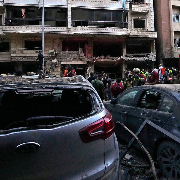 Räddningspersonal arbetar vid en skadad byggnad efter en explosion. I förgrunden syns två skadade bilar, med en av dem med trasig bakruta. Bakom dem är en skadad byggnad med flera öppningar och människor som evakuerar området. Räddningsfordon är synliga i bakgrunden.