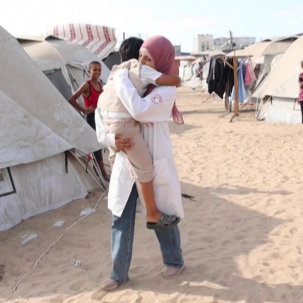 En sjukvårdare i Gaza bär ett barn genom ett flyktingläger med tält på båda sidor. Sandig mark syns mellan tälten, där flera personer är synliga i bakgrunden. Atmosfären tyder på en situation med humanitärt stöd.