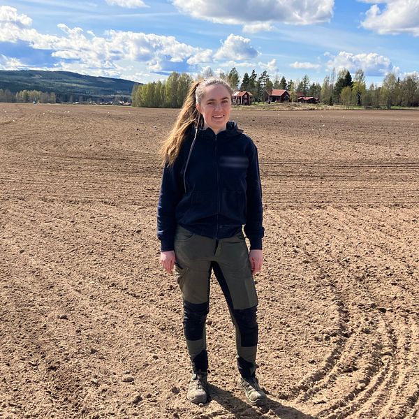 En kvinna står på en öppen åker under blå himmel med moln och skog i bakgrunden.