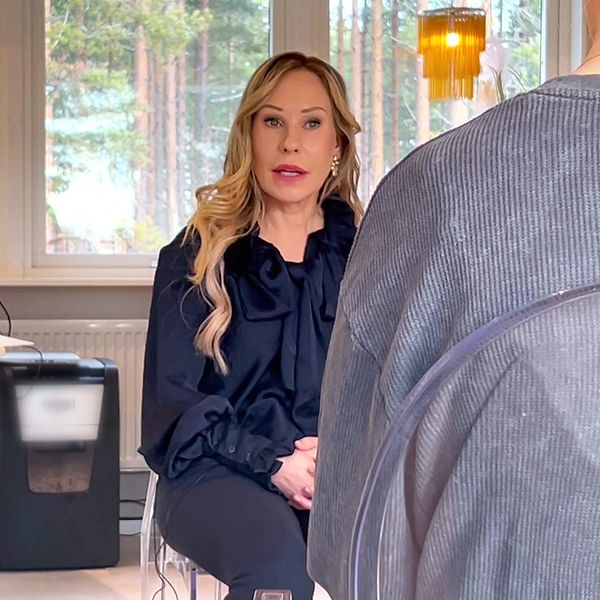 En blond kvinna sitter framåtlutad i en modern inomhusmiljö, med ett skrivbord i bakgrunden. Reportern sitter i en stol mittemot med ryggen mot kameran.