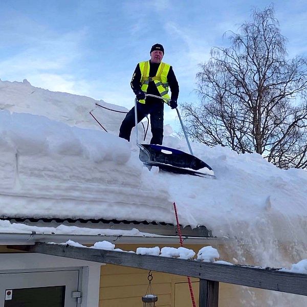 Man med varselväst står med en snösläde på ett villatak med snö.