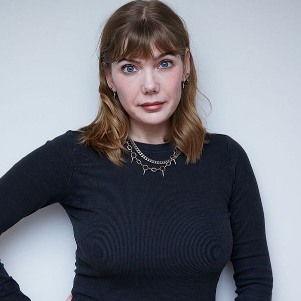 En person, Emily ”Modemassakern” Dahl, står med ena handen på höften, iförd en svart tröja och en kedjesmycke. Bakgrunden är ljus och neutral, vilket skapar fokus på personen. I övre vänstra hörnet finns en röd ikon med pratbubblor.