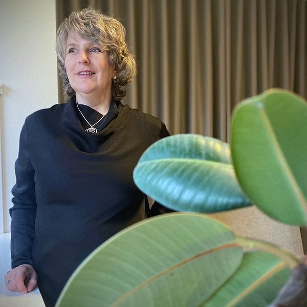 Pia Idhede Liljegren står i en inomhusmiljö iklädd en svart tröja, riktad mot kameran. I förgrunden syns gröna blad från en växt vilket ger en känsla av livfullhet i rummet. Bakgrunden består av ljusa väggar och en gardin.