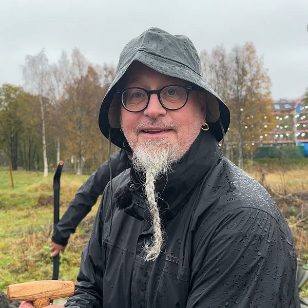 En person i regnkläder står i en fältmiljö, omgiven av träd och byggnader i bakgrunden. Det regnar och marken är blöt, medan personen håller i en träspade och fyller sandsäckar med sand.