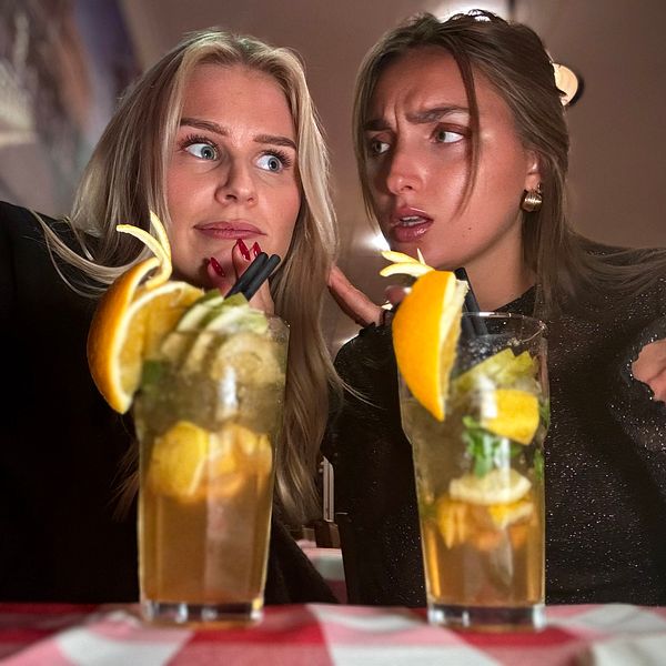 Två personer poserar med varsin drink på ett bord med röd-vitrutig duk. Drinkarna är dekorerade med citrusskivor och myntablad, vilket ger en livlig och uppfriskande känsla. I bakgrunden syns en suddig interiör av en restaurang.