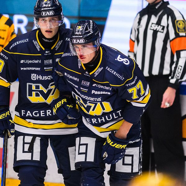 Två ishockeyspelare i matchtröjor från HV71 står tillsammans på isen. Den ena spelaren, Herman Hansson, ser ut att ha ont. I bakgrunden syns en domare i svart och vitt, och det är en energisk atmosfär i rinkens miljö.