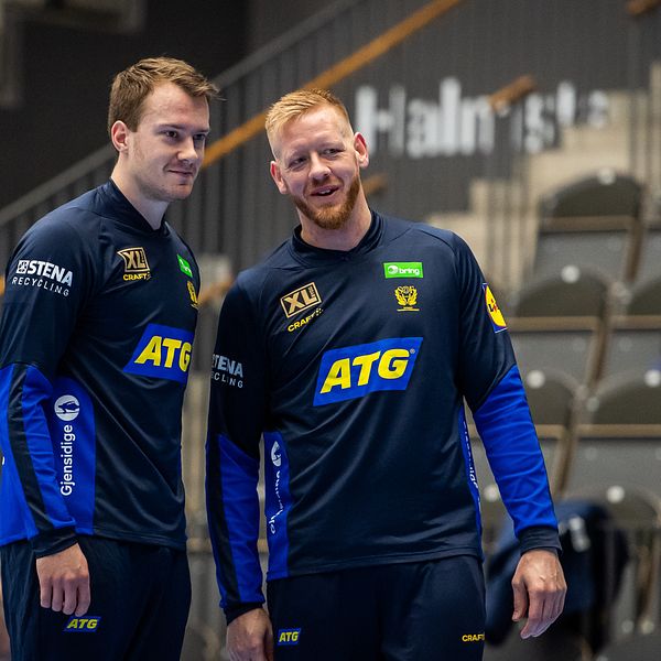 Två handbollsspelare i mörkblå träningskläder pratar på läktare inför match.