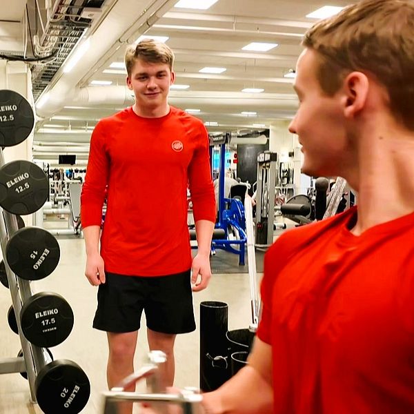 Två killar på ett gym, den ena drar i ett handtag för ryggträning medan hans kompis tittar på.