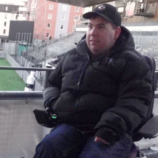 en man sitter i permobil iförd svart jacka och svart keps med ÖSKs emblem på. Bakom honom syns en läktare och en fotbollsplan.