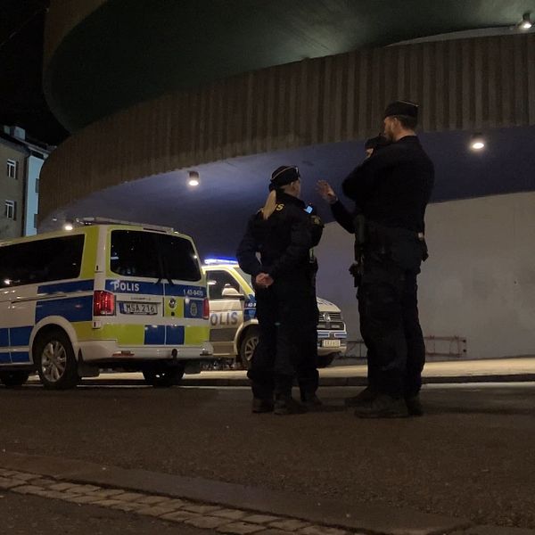 Två poliser står på en gata om natten i närheten av en polisbil, medan en byggnad med rundad struktur syns i bakgrunden.