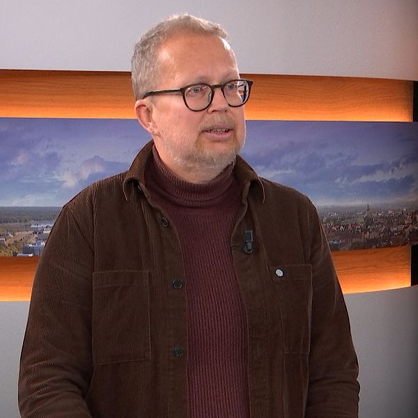 Personen är klädd i en mörkbrun jacka över en röd tröja och står framför en bakgrund med landskapsbilder, vilket tyder på en nyhetsstudio.