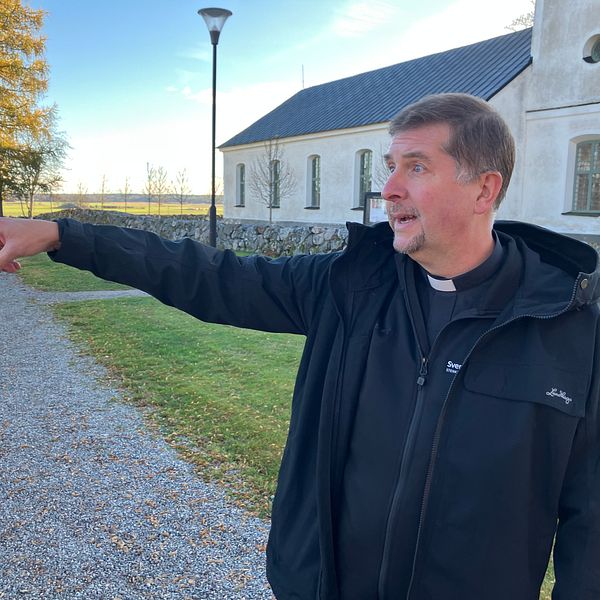 En kyrkoherde pekar med ena handen utanför Stenkvista kyrka. Det är en klar dag med ett par träd i bakgrunden och en lamppost syns i närheten. Gräset är grönt och välvårdat.