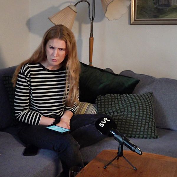 Kvinna sitter i soffa med mikrofon framför sig, anteckningar på soffan och mobil i handen.