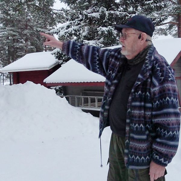 En person står i en snötäckt skog med en hand utsträckt och pekar mot en byggnad i bakgrunden. I närheten finns flera röda stugor omgivna av snötyngda träd. Det råder en kall och vintrig atmosfär.
