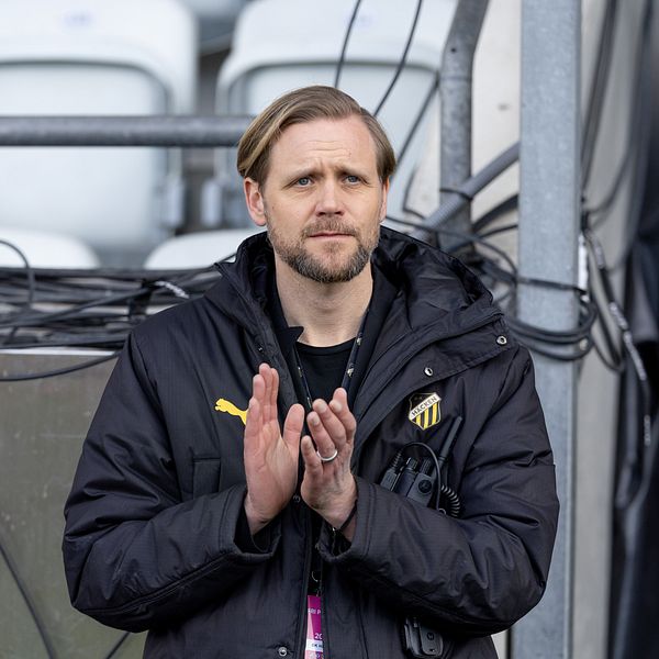Person i svart sportjacka applåderar vid sidan av läktare med tomma stolar i bakgrunden.