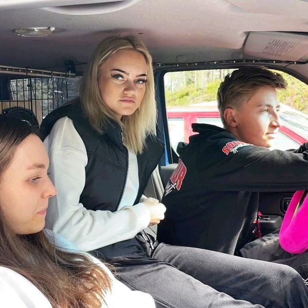 Tre ungdom sitter i en a-traktor – till vänster sitter en tjej med mörkt långt hår, i mitten av tjej med blont hår och vid ratten sitter en kortklippt kille. Det hänger wunderbaums, färggranna doftgranar, i fönstret runt backspegeln.