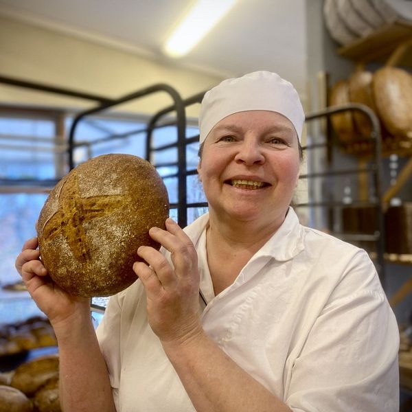 en glad kvinna i bagarekläder håller upp ett bröd. Tina Fernlund Järna