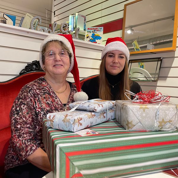Två personer sitter på en röd soffa, klädda i juliga tomteluvor. Framför dem finns en julklappar med olika inslagningar, inklusive en med röd och vit design och en annan med silverpapper. Bakgrunden har hyllor med olika föremål, vilket ger en festlig atmosfär.
