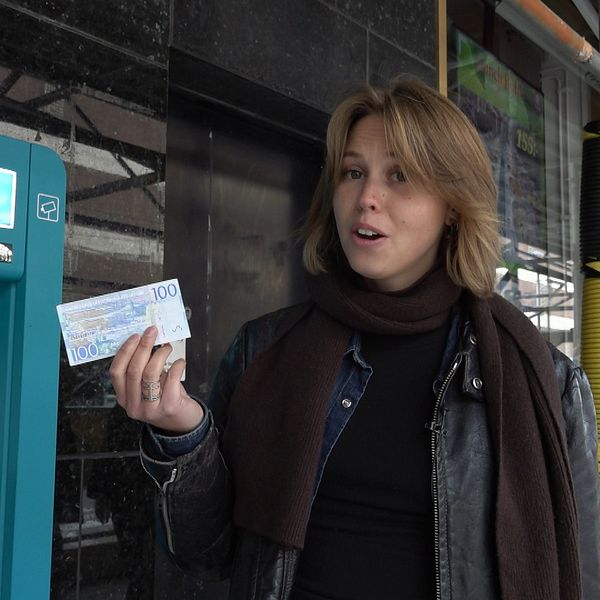 En person står vid en bankomat och håller upp en hundralapp. I bakgrunden syns en byggarbetsplats med rör och en glasruta.