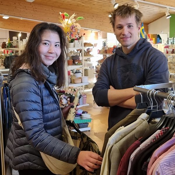 Två personer står i en butik med kläder och diverse inredning. Den första personen, klädd i en svart jacka, håller en väska och ser på kläder som hänger på en klädhängare. Den andra personen, i en blå tröja, står med armarna i kors och tittar på den andra personen. Butiken har ljusa väggar och en mängd olika varor syns i bakgrunden.