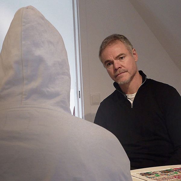 Två personer sitter mittemot varandra i ett inomhusmiljö. En person har på sig en vit huvtröja och är vänd bort från kameran, medan den andra personen, klädd i svart, sitter med fokus på den förra. Omkring dem finns en bordsyta med ett mönstrat föremål. Rummet har en modern inredning med ljusa väggar och en dörr i bakgrunden.