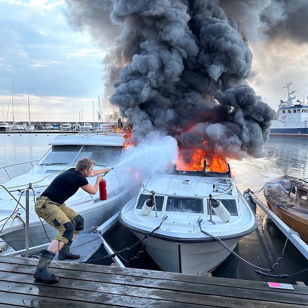 förtöjd motorbåt brinner kraftigt intill andra båtar, ihopkrupen man med brandsläckare försöker bekämpa lågorna från bryggan