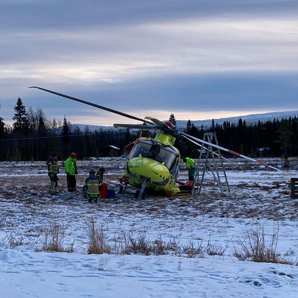 Räddningspersonal arbetar runt en helikopter som har landat på en snötäckt äng. Helikoptern är grön och orange, och snön omger området. Några personer är på väg att hjälpa en patient i medvetslös tillstånd på marken. I bakgrunden syns träd och en fjärran bergskedja under en molnig himmel.