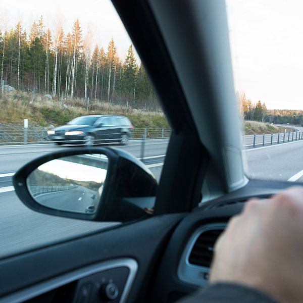 Vy från förarsätet i en bil som färdas på en motorväg, med en hand på ratten. En annan bil syns som kör i motsatt körfält. Tallskog och bar mark skymtas längs vägkanten.