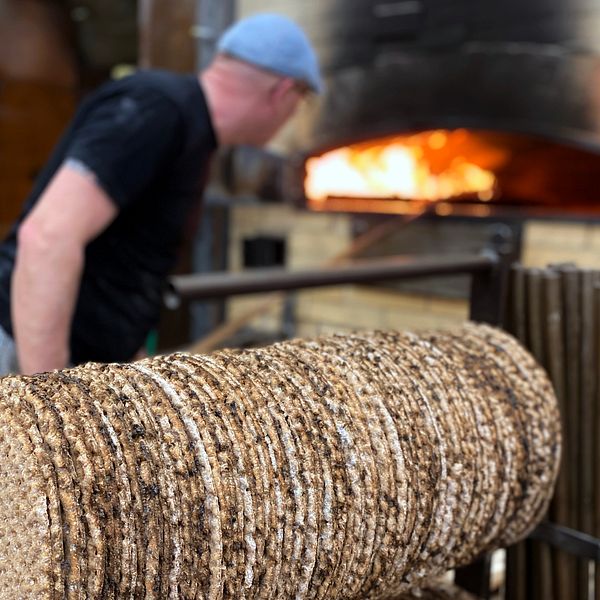 Stora Skedvi, knäckebrödsfabrik. Knäckebröd hänger på tork i förgrunden. I bakgrunden står en bagare och lägger in bröd i en vedeldad stenugn.