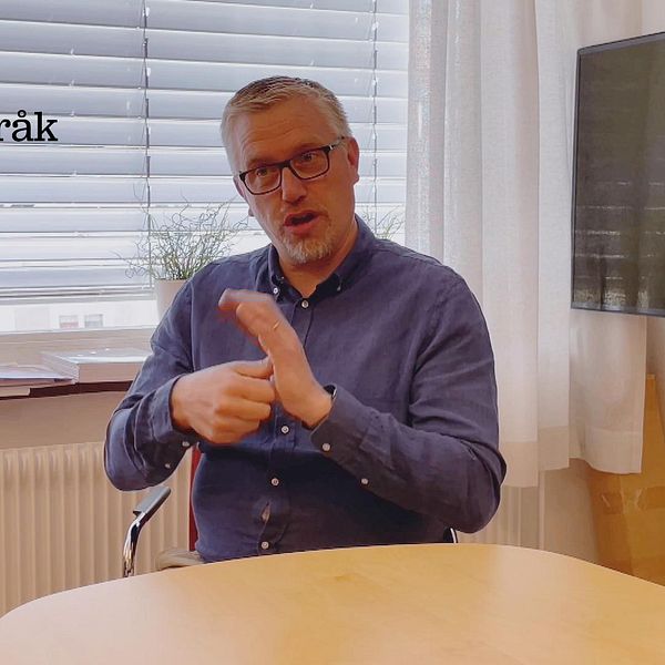 Roger Holmström har en blå skjorta på sig och sitter vid ett bord. Bakom honom finns ett fönster och till höger finns en tv.