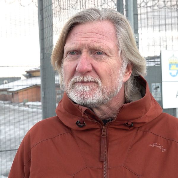 En person står framför ett stängsel på en anstalt, klädd i en röd jacka. I bakgrunden syns en byggnad samt en skylt med texten ”Kriminalvården Anstalten Umeå”. Det är en kall och klar dag, vilket framgår av den snöiga omgivningen.