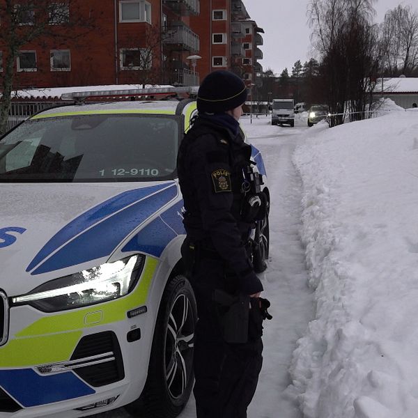 En polisbil står parkerad på en snöig gata framför en flerbostadsbyggnad. En polis i uniform står bredvid bilen och tittar bortåt längs gatan. Det är vinter och landskapet är täckt av snö.