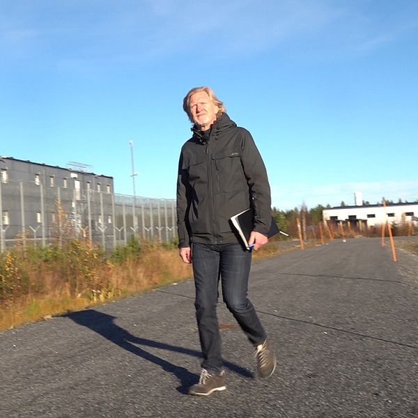 En person går längs en asfaltväg som leder förbi en inhägnad byggnad. Omgivningen är grön och solig, med en klar blå himmel. Längs vägkanten står orangea markörer.