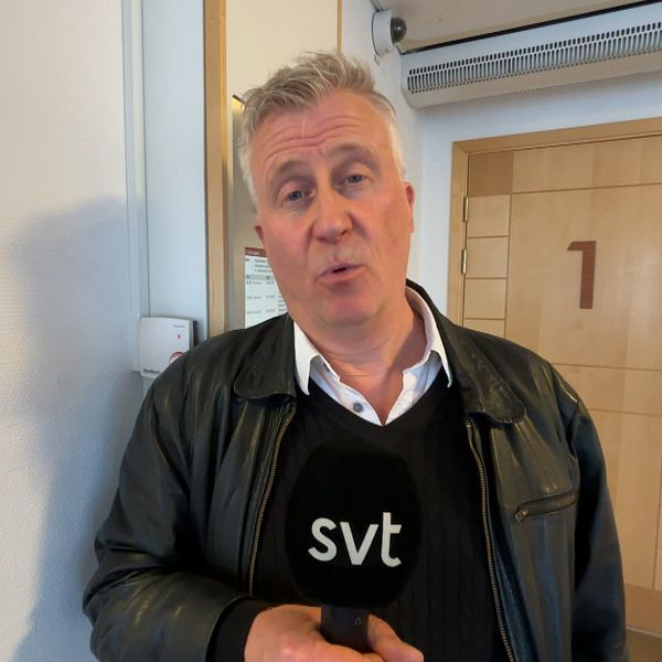 Person med grått hår håller en SVT-mikrofon i en korridor utanför en dörr märkt med en etta.