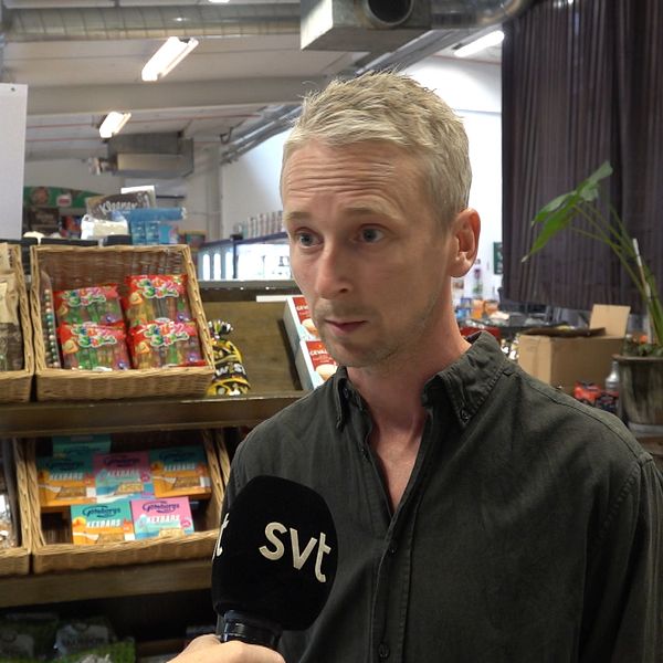En man med blont hår och skjorta står i vad som ser ut att vara en liten matbutik.