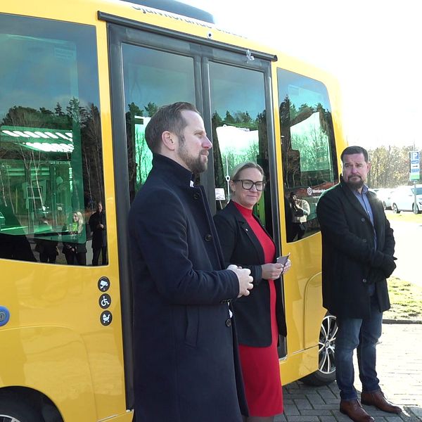 Tre personer står framför en gul självkörande buss. Två av dem är klädda i mörka rockar, medan den tredje bär en röd klänning. Bussen är parkerad på en asfaltväg, med en ljus bakgrund av träd och byggnader.