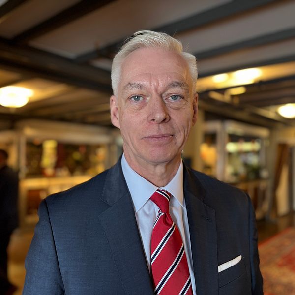 Man i kostym och röd slips står i receptionen. En annan person syns i bakgrunden.