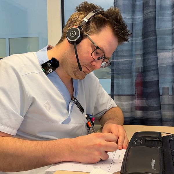 En sjuksköterskeklädd man sitter vid ett skrivbord och skriver för hand. På huvudet har han ett headset.