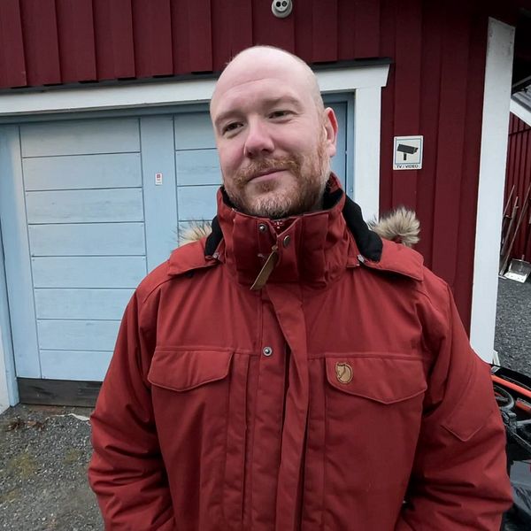 En person i en röd jacka står framför en röd byggnad med blå garageportar. På vänster sida finns en lampa som lyser, och i bakgrunden syns en uppfart med grus samt en del av en staket inramande området.