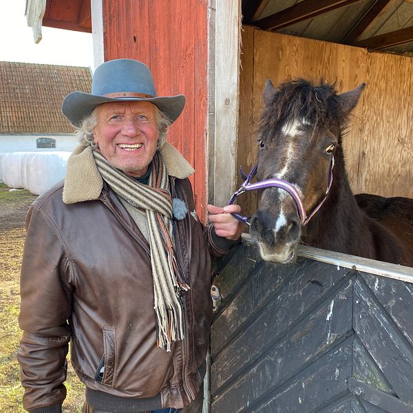 En person i läderjacka och scarf står vid en hage och interagerar med en svart häst som sträcker sig fram genom stallets öppning. I bakgrunden syns en lada och andra delar av gården.