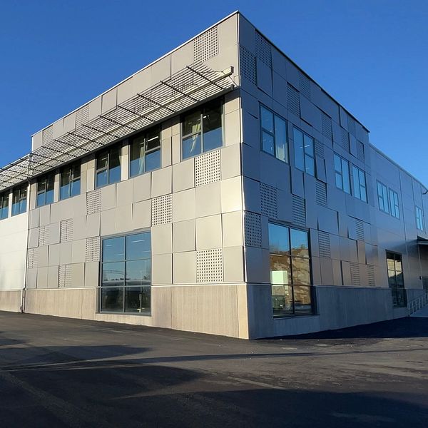 Industribyggnad i solljus med moderna fönster och platt tak, belägen på en asfalterad yta under en klarblå himmel.