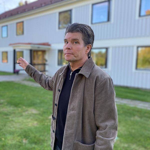 Mattias Fjellström ny chef på Sis ungdoms Vemyra står utomhus och gestikulerar med en arm, framför en byggnad med flera fönster. Området runt dem är grönt med gräsmatta och en stenlagd gång. Byggnaden har en ljus färg och ett rött tak, vilket ger en kontrast till den omgivande naturen.