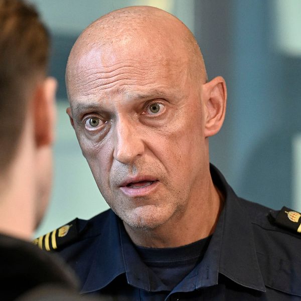 Två personer samtalar, en i polisuniform och en med ryggsäck, inomhus vid grönt draperi.