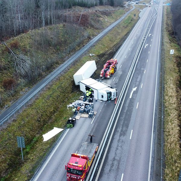 Lastbil som har vält på en motorväg, med räddningsfordon på plats för att hantera situationen. Krossat gods ligger utspritt på vägbanan och det omgivande området är delvis skogbeklätt.