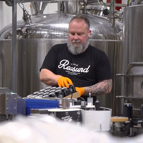 En man med skägg och svart T-shirt står och packar ölburkar inne på ett bryggeri.