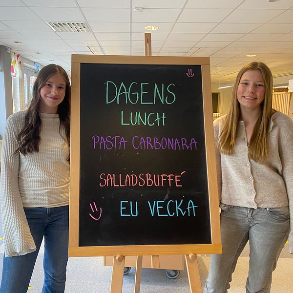 Två personer står vid en meny-tavla där det står ”Dagens lunch: Pasta carbonara, salladsbuffé, EU-vecka”.