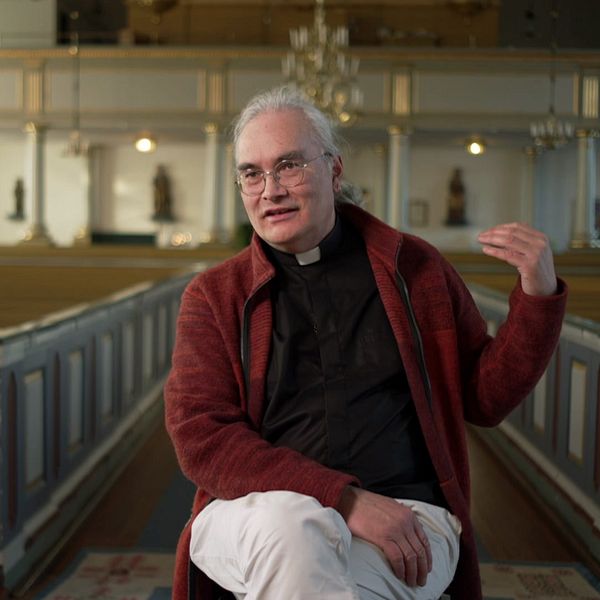 Prästen Oliver Li i Forsa kyrka med tomma bänkar och ljuskrona i bakgrunden.