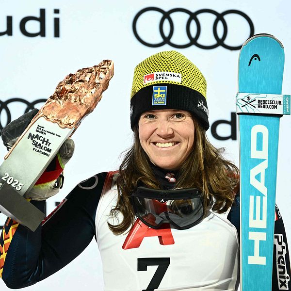 En pristagare i skidsport håller upp en trofé och en skida, klädd i en tävlingsdräkt med nummer 7. I bakgrunden syns flera Audi-logotyper som visar på sponsorernas närvaro vid evenemanget.