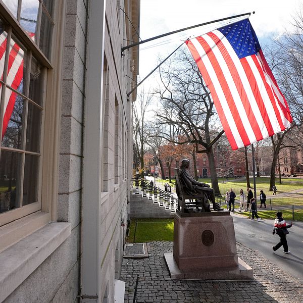 USA-flagga utanför universitetsbyggnad, studenter promenerar på gångväg i bakgrunden.